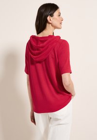 Röd kortärmad topp med draperad huva, med en avslappnad passform och mjuk textur. Designen är enkel utan synliga mönster eller detaljer.