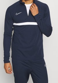 Granatowa sportowa bluzka z długim rękawem z białym poziomym paskiem, suwakiem przy szyi i logo Nike. Materiał wydaje się gładki i rozciągliwy dla komfortu.