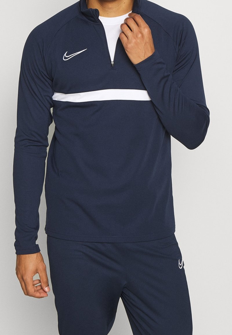 Granatowa sportowa bluzka z długim rękawem z białym poziomym paskiem, suwakiem przy szyi i logo Nike. Materiał wydaje się gładki i rozciągliwy dla komfortu.