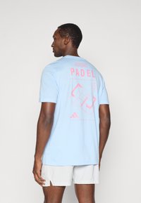 Jasnoniebieska bawełniana koszulka z różowym nadrukiem "PADEL" na plecach, przedstawiająca wzór boiska i logo Adidas na dole.