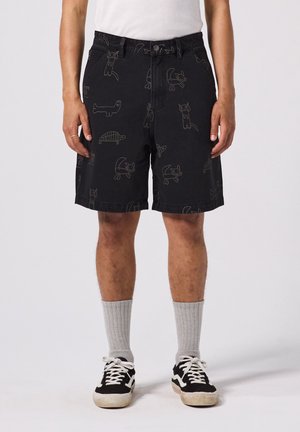 Man draagt zwarte korte broek met cartoon dierafdrukken, wit hemd, grijze geribbelde sokken en zwart-witte sneakers, staand tegen een effen achtergrond.