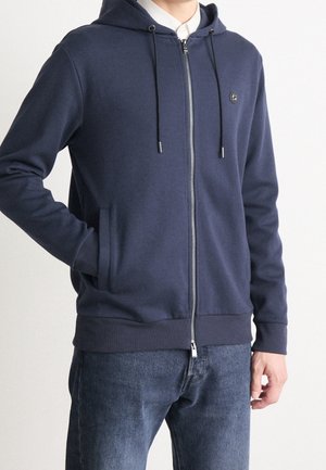 Homme portant un sweat à capuche zippé bleu marine avec cordons, par-dessus un t-shirt blanc, associé à un jean bleu, main dans la poche sur fond uni.