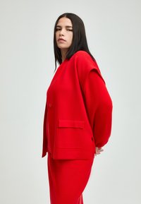 Roter übergroßer Cardigan mit lockerer Passform, ausgestattet mit einem einzigen Knopf, vorderen Taschen und einem strukturierten Strick. Kombiniert mit einer passenden Hose.