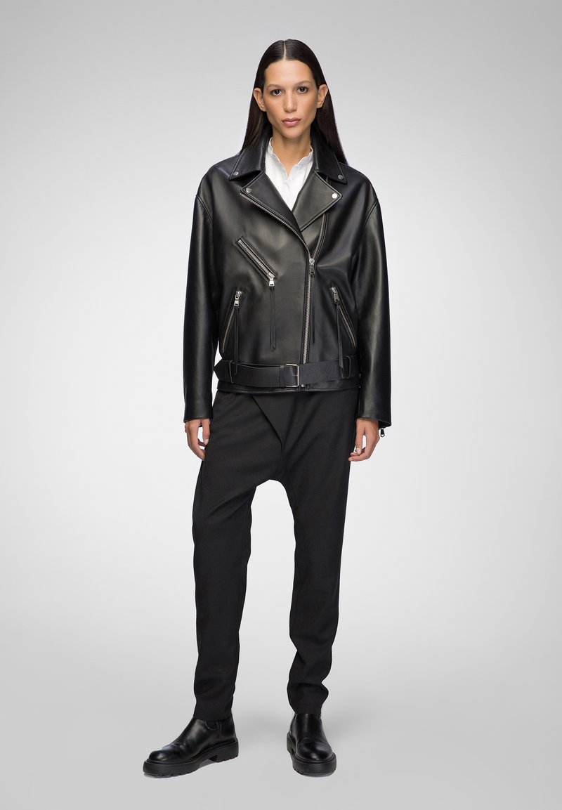 Schwarze Leder-Bikerjacke mit silbernen Reißverschlüssen, einem Gürtel und asymmetrischer Vorderseite. Kombiniert mit lockeren schwarzen Hosen und Stiefeletten.