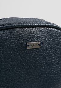 Tmavě modrý batoh s texturou, hladkým povrchem a stříbrnou štítkem s logem Roxy, přišitým tmavě modrým stehem. Zaoblený tvar se zipovým zapínáním.