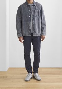 Giacca di denim grigia con davanti con bottoni e tasca sul petto, abbinata a pantaloni blu navy e sneaker grigio chiaro su un pavimento in legno.