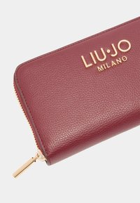 Bordo odinė piniginė su auksine įspausta logo "LIU·JO MILANO" ir auksinės spalvos užtrauktuku, pasižyminti stilingu dizainu ir tekstūruotu paviršiumi.