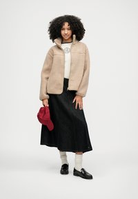 Beige fleecevest, wit shirt, lange zwarte denim rok, rode pet, zwarte loafers en crème sokken. Natuurlijk, krullend haar. Eenvoudig kledingontwerp.