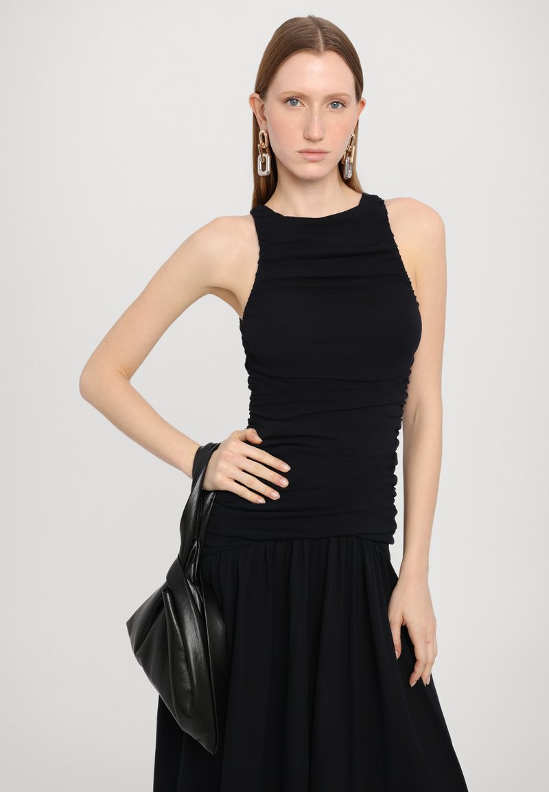 Schwarzes, eng anliegendes Tanktop mit geraffter Textur, kombiniert mit einem fließenden schwarzen Rock. Das Model hält eine schwarze Ledertasche und trägt große metallische Ohrringe.