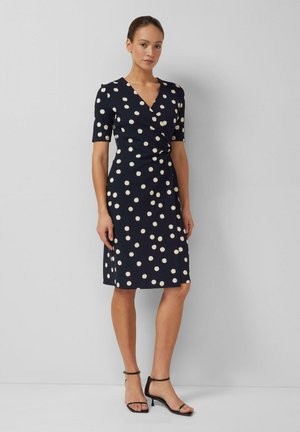 Frau steht vor einer grauen Wand, trägt ein knielanges marineblaues Kleid mit weißen Punkten und schwarze Knöchelriemen-High Heels.