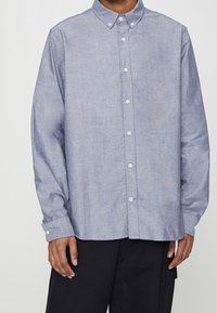 Homme portant une chemise boutonnée bleu clair à manches longues et un pantalon noir, debout devant un fond uni.