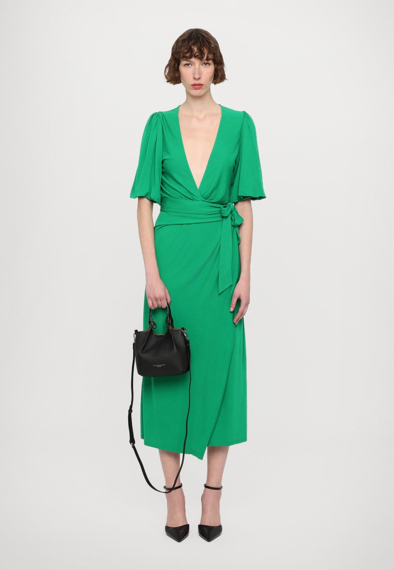 Green wrap dress with short puff sleeves and a deep V-neck, paired with a small black handbag and black high-heeled shoes.