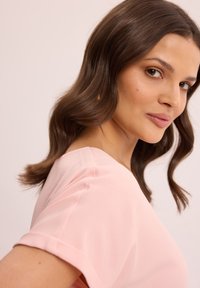 Młoda kobieta z falującymi brązowymi włosami, ubrana w jasnoróżową bluzkę z krótkim rękawem, patrząca przez ramię na tle jednolitego jasnego tła.