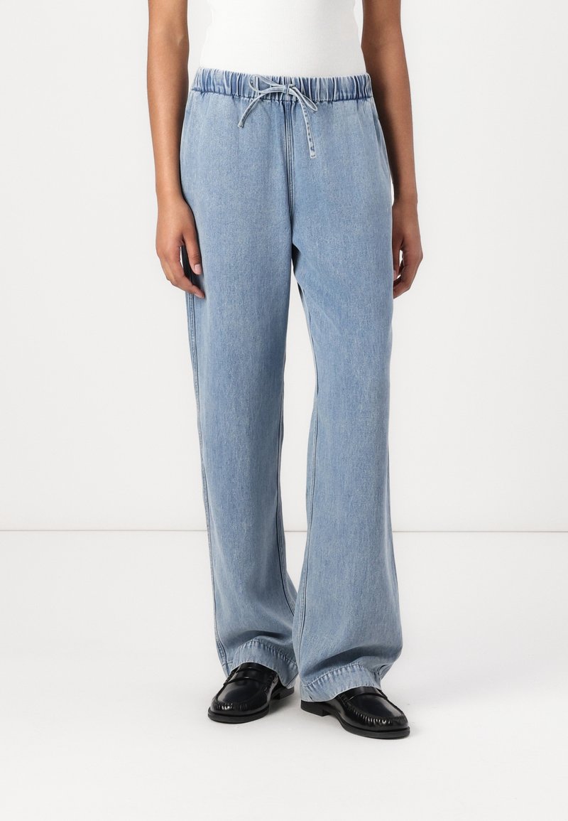 Personne portant un pantalon en denim bleu clair à jambes larges avec taille élastique et cordon, associé à des mocassins noirs.