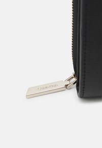 Black leather wallet with gold-toned zipper pull featuring a rectangular tag embossed with "Calvin Klein." Textured surface and sleek design.