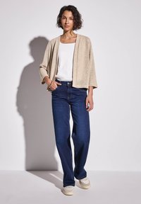 Beige öppenstickad kofta, vit topp och mörkblå vida jeans. Modellen bär ljusa sneakers och står mot en neutral bakgrund.