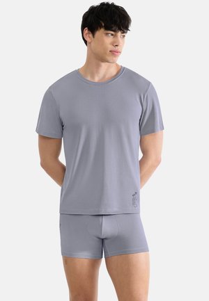 Young man with dark hair wearing a matching light gray t-shirt and shorts set, standing with hands behind back and looking to the side.