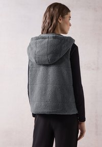 Gilet in fleece grigio con un motivo trapuntato e cappuccio, caratterizzato da una texture morbida e design senza maniche, indossato sopra una maglia nera a maniche lunghe.
