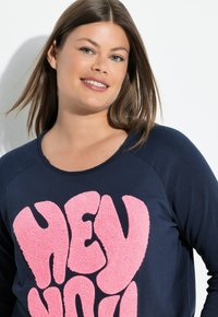 Sudadera de manga larga azul marino con un gráfico esponjoso en rosa que dice "HEY YOU" en el frente. Cuello redondo, tela suave, corte relajado.
