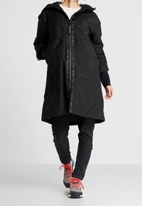 Abrigo negro impermeable con capucha y cierre de cremallera. Cuenta con grandes bolsillos y un diseño hasta la cintura. Combinado con pantalones negros y zapatos coloridos.