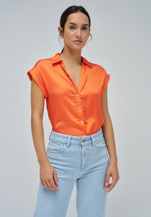 Blusa de satén naranja de manga corta con cuello, botones en la parte delantera y mangas ligeramente enrolladas, combinada con jeans de tiro alto de color azul claro.