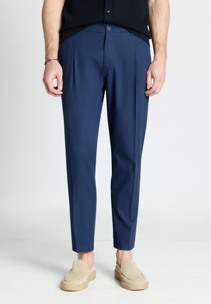 Homme portant un pantalon slim bleu marine et des chaussures beiges sans lacets, debout devant un fond uni.