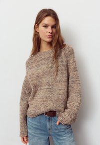 Neulepusero beigen ja kerman sävyissä, löysällä istuvuudella, jossa on resorireunainen pääntie ja leveät hihat, yhdistetty vaaleansinisiin farkkuihin.