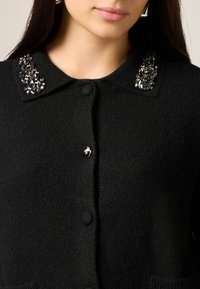 Cardigan nero in tessuto strutturato, con chiusura a bottoni e colletto decorato con pietre preziose trasparenti e nere.