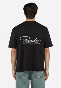Black short-sleeve t-shirt with a relaxed fit. Back features white cursive text "Pegador" and "ALL RIGHTS RESERVED." Soft cotton material.