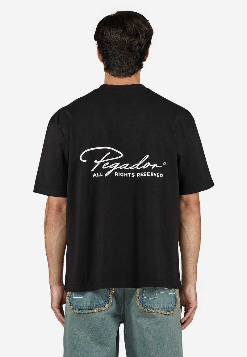 Black short-sleeve t-shirt with a relaxed fit. Back features white cursive text "Pegador" and "ALL RIGHTS RESERVED." Soft cotton material.