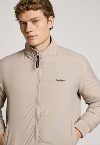 Beige vadderad jacka med hög krage, framtida dragkedja och svart logotyp på bröstet. Har ribbade ärmslut och två sidofickor.