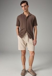 Brun kortærmet skjorte med knapper og teksturerede striber, parret med beige teksturerede shorts. Modellen har beige sokker og brune slip-on sko på.