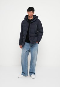 Navy puffer jacket with a hood, featuring a front zipper, two side pockets, and a left chest pocket; worn over a black shirt and blue jeans.