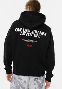 Osoba v čierneho mikiny s kapucňou, na ktorej je na chrbte potlač "POSLEDNÁ ZÁHADNÁ DOBRODRUŽSTVO" a text "Stranger Things", v kombinácii so svetlošedými džínsami.