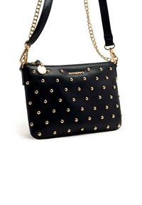 Bolso de mano de cuero negro con tachuelas doradas dispuestas en un patrón de puntos, que cuenta con una cadena y una correa, además de un charm redondo en el lateral.