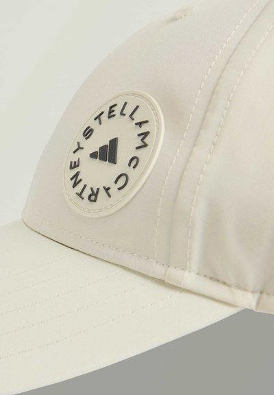 Beige baseball cap with a curved brim, featuring a round, black logo patch with white text and design accents. Smooth fabric texture.