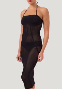 Donna che indossa un vestito nero trasparente in rete con spalline a collo alto sopra biancheria nera, in piedi contro uno sfondo chiaro e semplice.