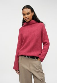 Pārāk liels, ribots adījuma džemperis tumši rozā krāsā ar augstu apkakli. Apvienots ar bēšiem biksēm, kas atspoguļo brīvu piegriezumu un maigu tekstūru.