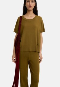 Zielono-oliwkowy sweter z szerokim dekoltem i krótkimi rękawami, w zestawieniu z pasującymi spodniami. Czerwona torba z frędzlami wisi na ramieniu.