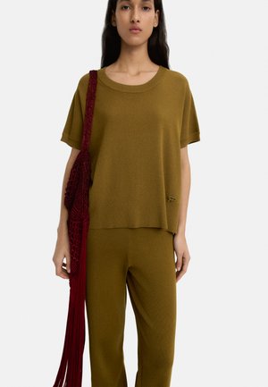 Zielono-oliwkowy sweter z szerokim dekoltem i krótkimi rękawami, w zestawieniu z pasującymi spodniami. Czerwona torba z frędzlami wisi na ramieniu.