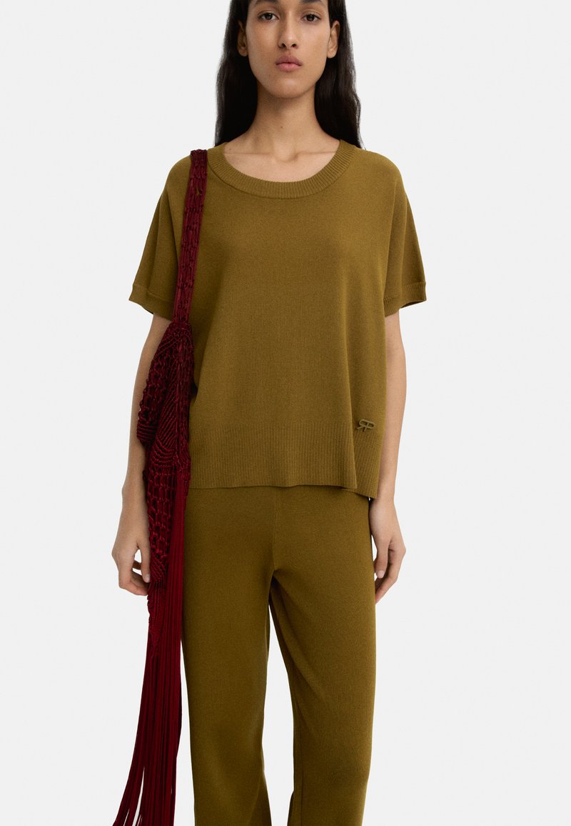 Zielono-oliwkowy sweter z szerokim dekoltem i krótkimi rękawami, w zestawieniu z pasującymi spodniami. Czerwona torba z frędzlami wisi na ramieniu.