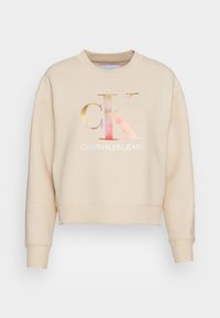 Zkrácená béžová mikina vyrobená z jemné látky, která má velké gradientní kovové logo "CK" a text "CALVIN KLEIN JEANS" pod ním.