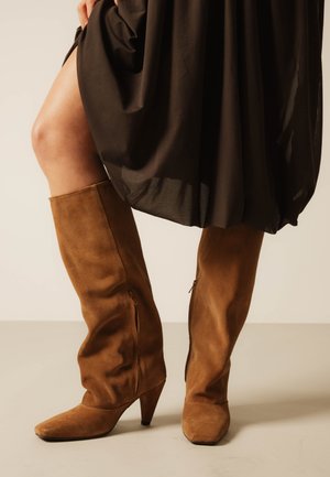 Femeie purtând cizme maro din piele întoarsă până la genunchi, cu vârful ascuțit și tocuri din lemn, purtate cu o rochie neagră care ridică ușor tivul.