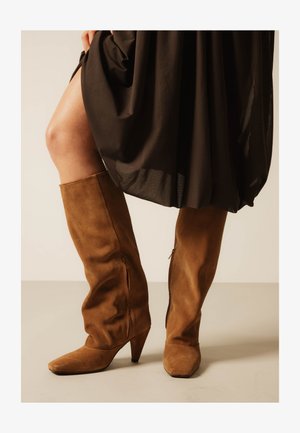 Woman wearing knee-high brown suede boots with pointed toes and wooden heels, paired with a black dress lifting the hem slightly.