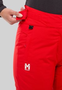 Pantalon de ski rembourré rouge avec ceinture velcro réglable, poche avant et logo Millet. Le matériau semble durable et résistant à l'eau.