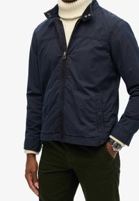 Granatowa kurtka wykonana z lekkiego materiału, z zamkiem z przodu, kołnierzem i kieszeniami po bokach. Noszona na kremowym swetrze z golfem.