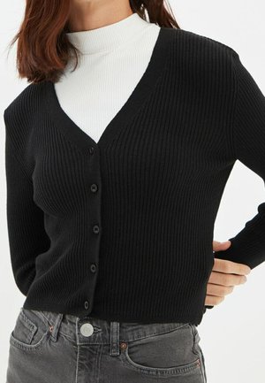 Cardigan noir à côtes avec un col en V, doté d'une fermeture à boutons et de manches longues, porté sur un haut blanc à col montant. Tissu texturé.