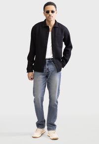 Black textured button-up shirt over a white tank top, paired with light blue jeans and beige sneakers. Model wears sunglasses.