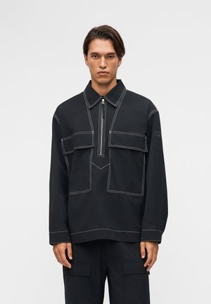 Mand iført en sort langærmet skjorte med hvide syninger, en kort lynlås foran, to brystlommer og matchende sorte bukser.