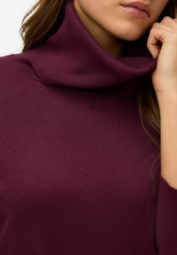 Maglione a collo alto borgogna realizzato in morbido tessuto a coste, con un colletto alto e una superficie liscia, parzialmente visibile su un modello.